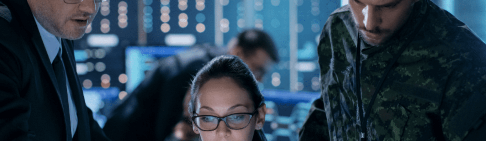 Girl in combat suit sitting at computer with two men behind working with her